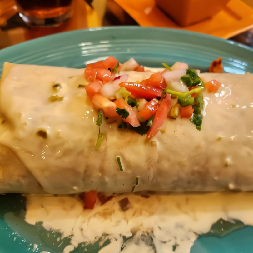 A large burrito covered in white sauce and topped with pico de gallo on a blue plate.