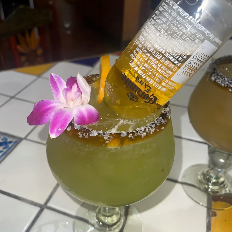 A green El Rodeo margarita with a Corona bottle, orange slice, and purple orchid on a tiled table.